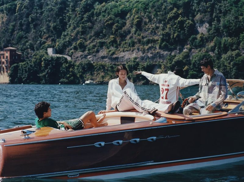Three people in casual wear sit on a wooden speedboat on a lake surrounded by lush greenery and a building in the distance.