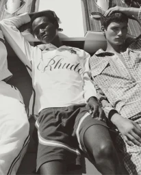 Black and white editorial fashion photograph of male models reclining near a boat for Rhude High Summer 2025.