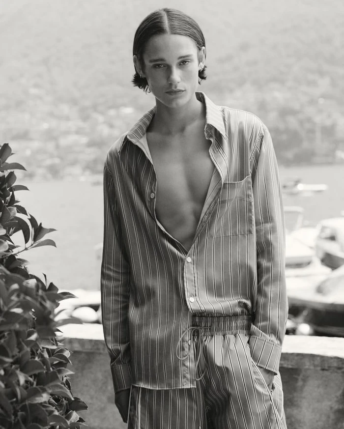 Monochrome portrait of a young male model wearing a striped shirt and matching trousers outdoors by water.