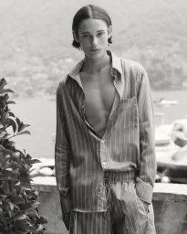Monochrome portrait of a young male model wearing a striped shirt and matching trousers outdoors by water.