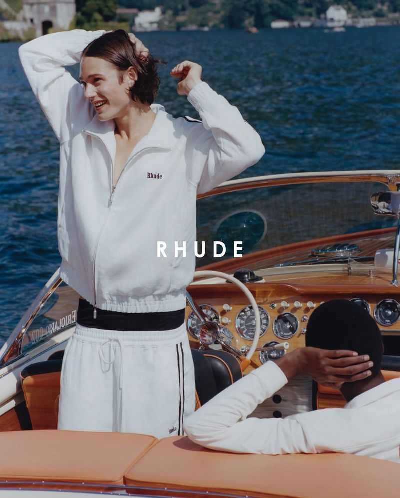 Model wearing a white zip-up jacket and white pants standing on a wooden boat on the water.