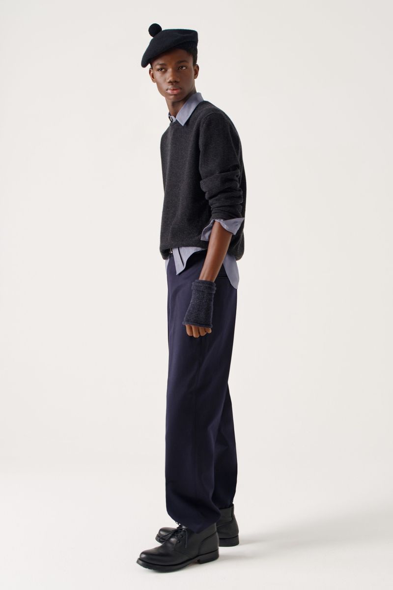 Dark-skinned man in a beret, sweater, and trousers strikes a pose in a fashion editorial shot in a brightly lit photo studio setting.