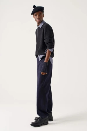 Dark-skinned man in a beret, sweater, and trousers strikes a pose in a fashion editorial shot in a brightly lit photo studio setting.
