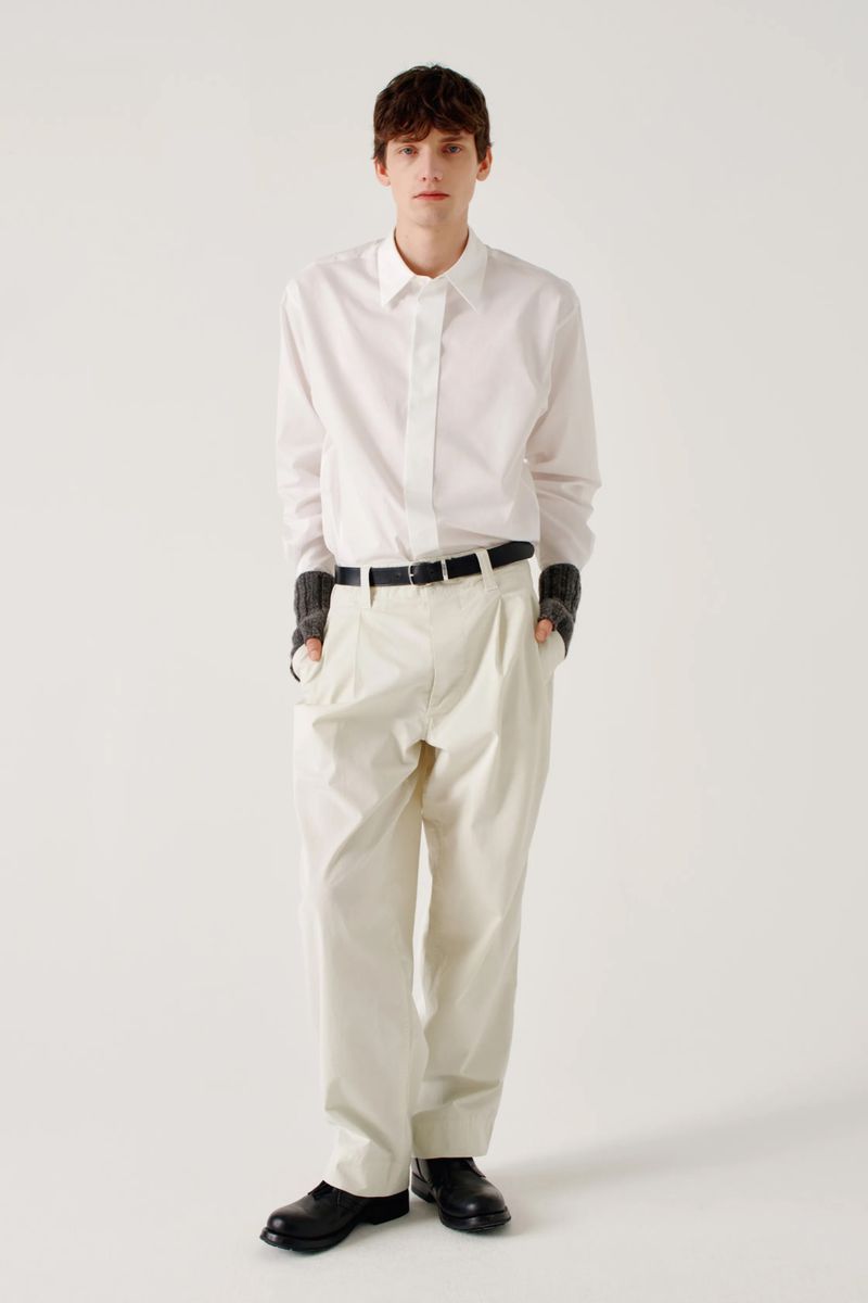 Full shot of a young adult man in a white button-down shirt and beige pants with black belt and shoes posing in a bright photo studio.