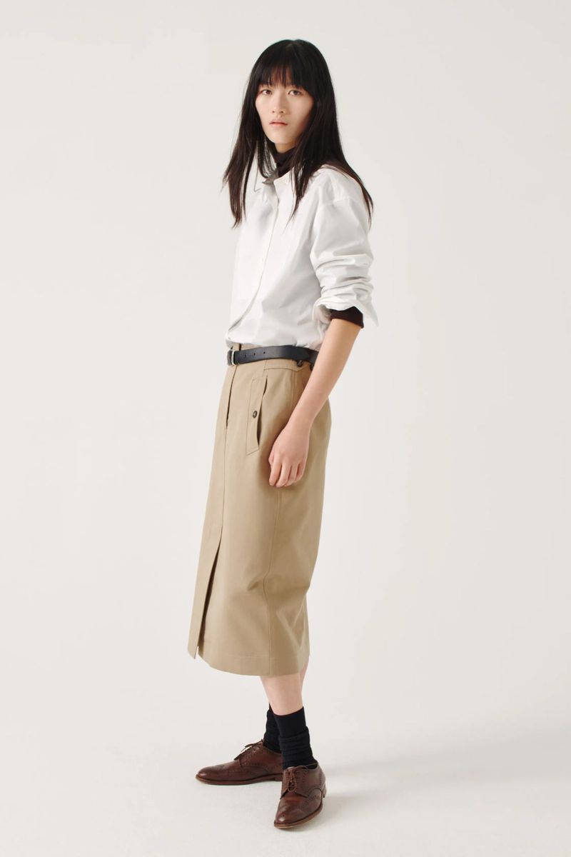 Full shot of a woman with dark hair in a white button-down shirt and tan skirt, wearing brown leather shoes against a white background.