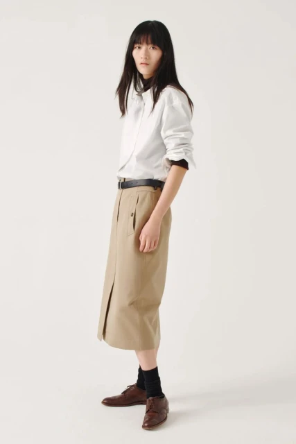 Full shot of a woman with dark hair in a white button-down shirt and tan skirt, wearing brown leather shoes against a white background.