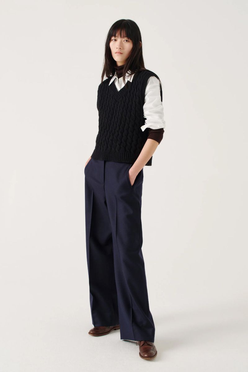 Full-length shot of a young Asian woman in a fashion pose, wearing a cable-knit sweater vest, white shirt, navy pants, and brown leather shoes.