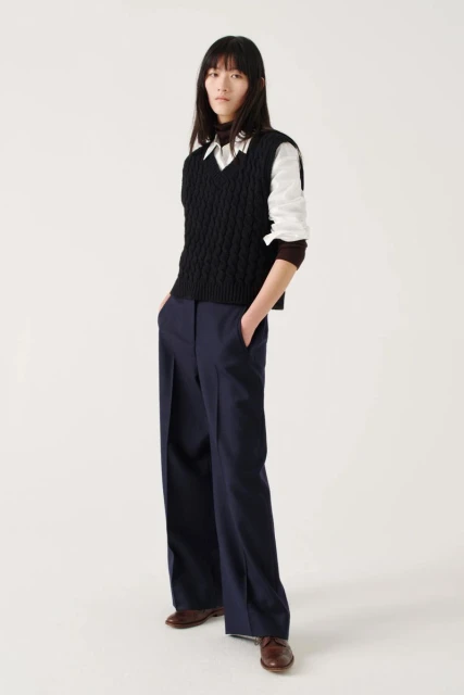 Full-length shot of a young Asian woman in a fashion pose, wearing a cable-knit sweater vest, white shirt, navy pants, and brown leather shoes.