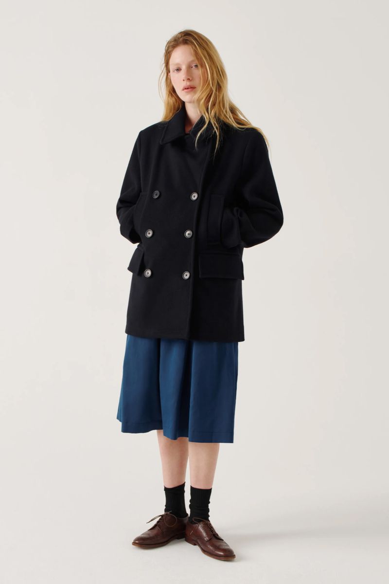 Full-length shot of a woman with blonde hair in a navy double-breasted jacket, blue midi skirt, black socks, and brown leather dress shoes in a studio.