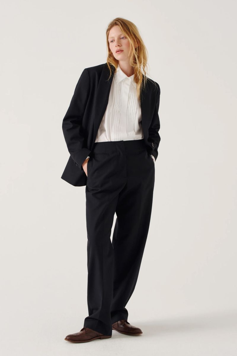 A woman with wavy blonde hair wearing a black suit and white pleated shirt standing against a plain white background.