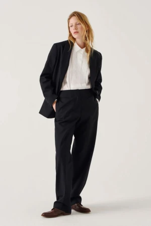 A woman with wavy blonde hair wearing a black suit and white pleated shirt standing against a plain white background.
