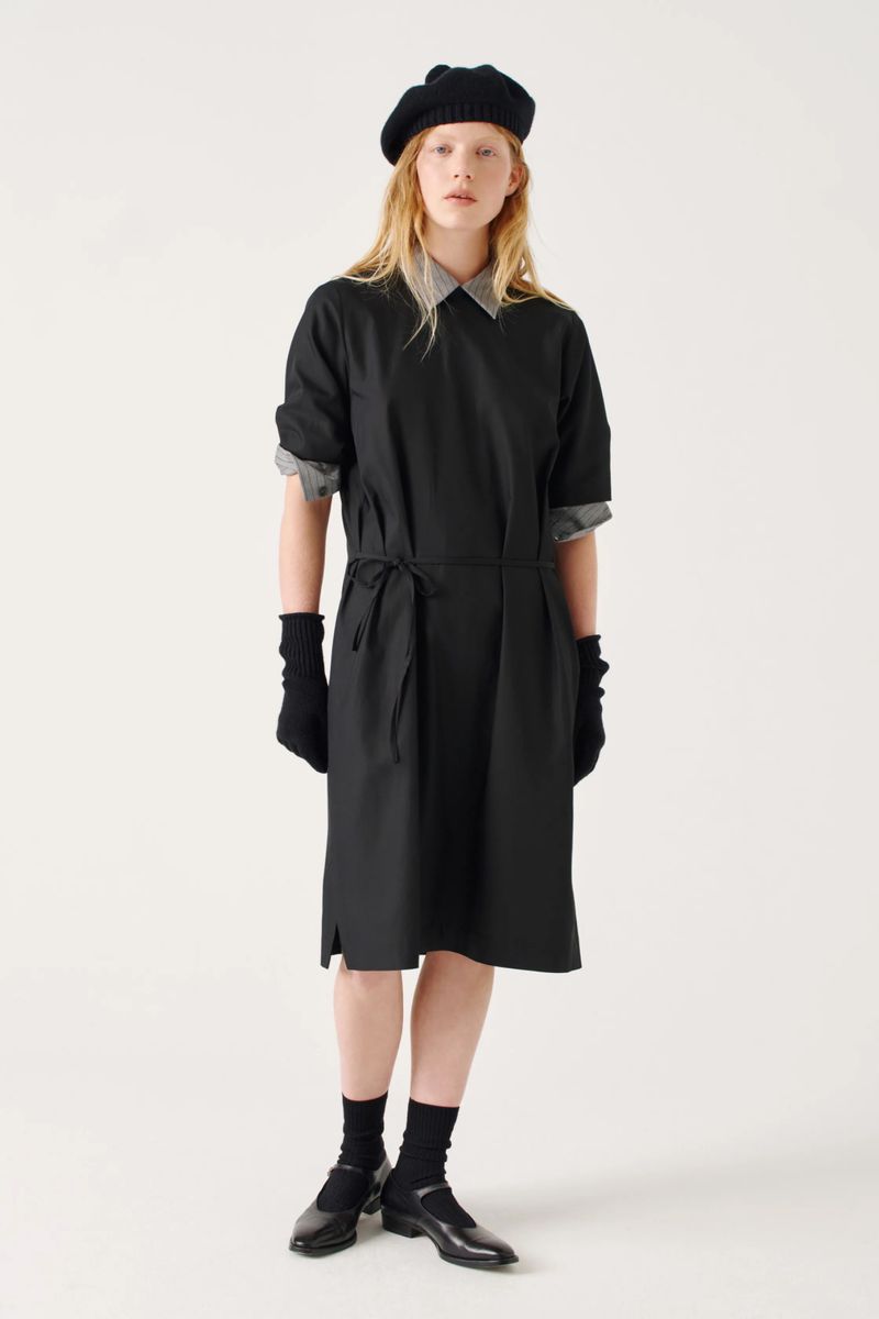 A young woman stands in a black dress, tights, and Mary Jane shoes, wearing a beret and gloves against a solid white background.