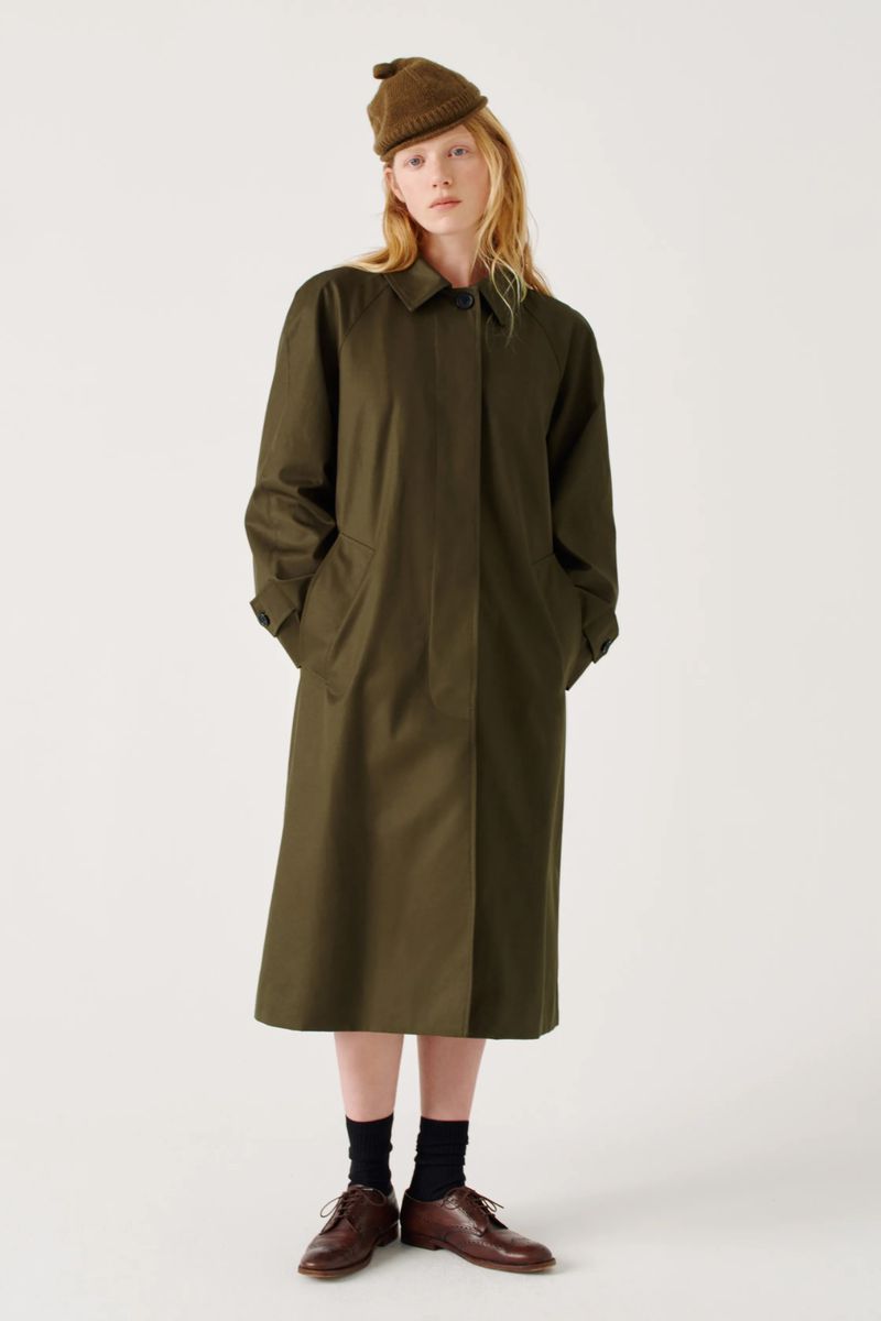 Full-length shot of a fair-skinned woman in an olive-green trench coat and knit hat, standing against a plain white backdrop in a studio setting.