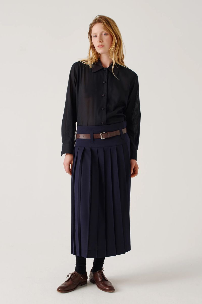 Standing woman in black button-down shirt, pleated midi skirt, and brown leather shoes poses in a bright studio setting.