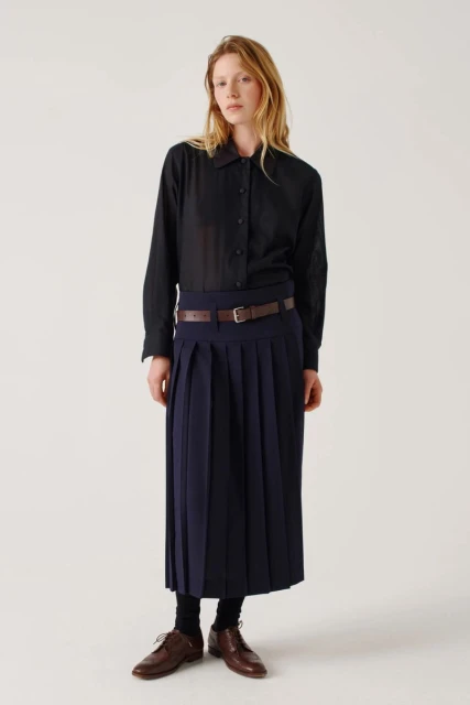 Standing woman in black button-down shirt, pleated midi skirt, and brown leather shoes poses in a bright studio setting.