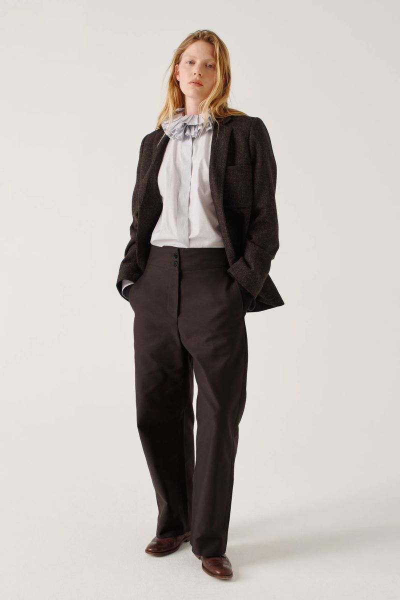 Full-length studio shot of a person with red hair, wearing a dark blazer, pants, and brown shoes, posing against a white backdrop.