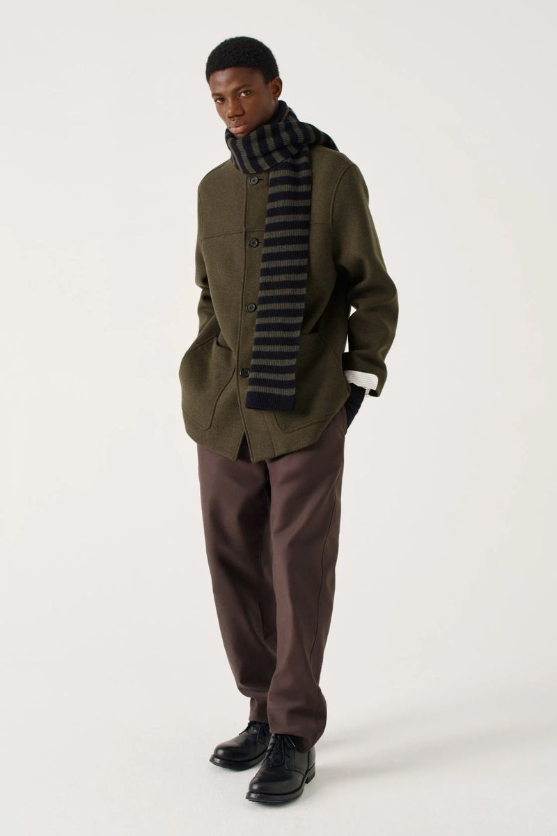 An adult Black man poses in a studio wearing an olive jacket, brown trousers, black shoes, and a black and gray striped scarf.