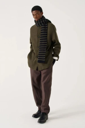 An adult Black man poses in a studio wearing an olive jacket, brown trousers, black shoes, and a black and gray striped scarf.