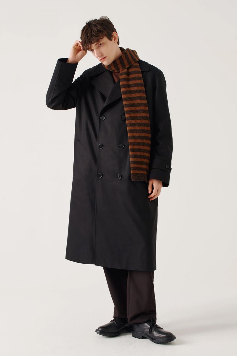 Male fashion model poses in a black trench coat and striped scarf against a white background in a studio shot. 