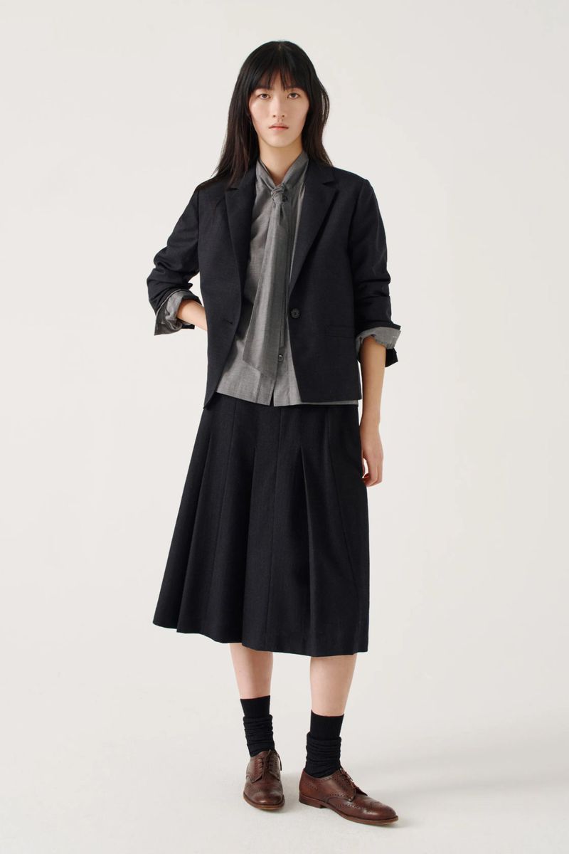 Model wearing a black blazer over a grey patterned shirt and pleated black skirt against a white background.