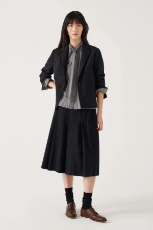Model wearing a black blazer over a grey patterned shirt and pleated black skirt against a white background.