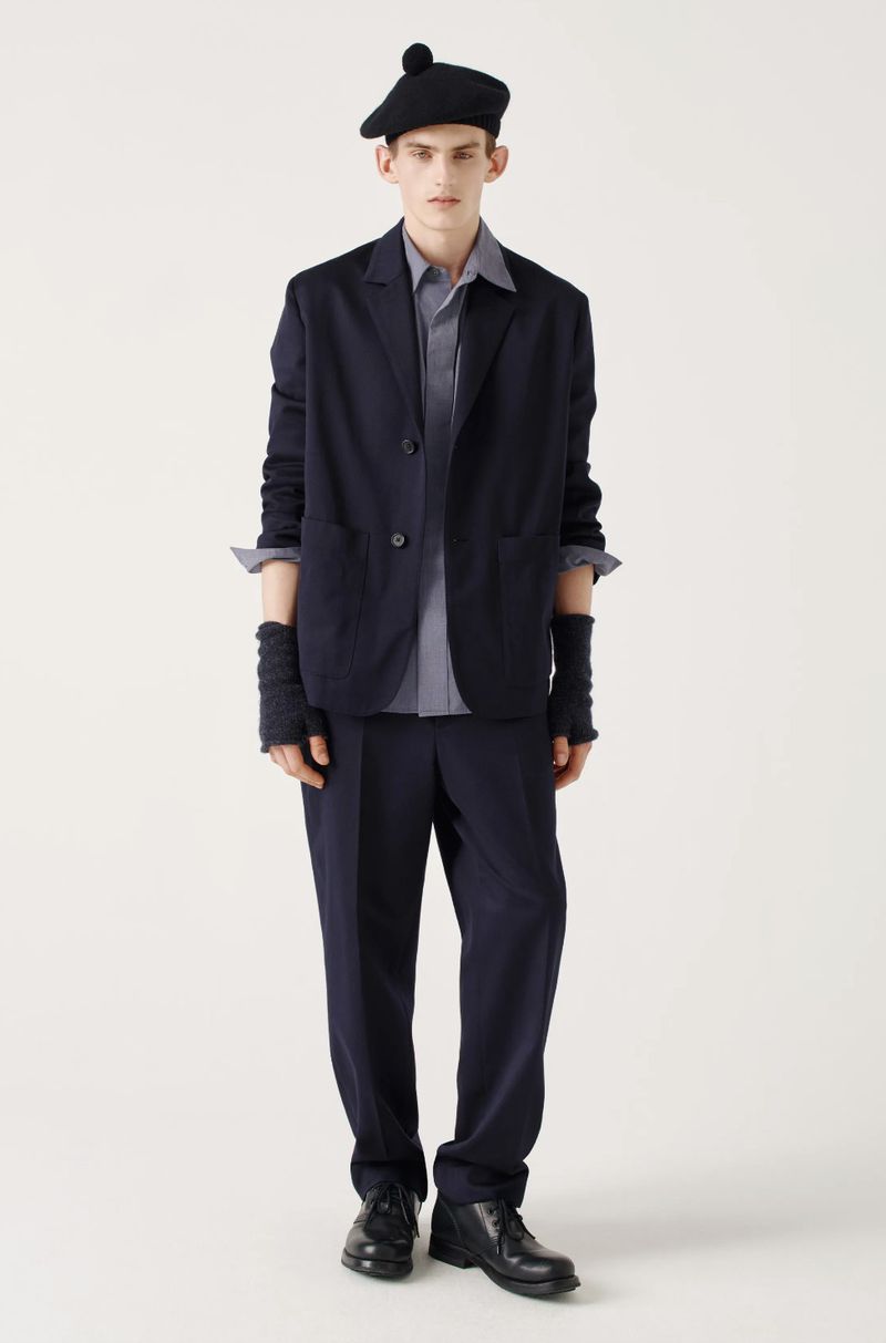 Full-length studio shot of a young adult man in a blazer, shirt, trousers, beret, fingerless gloves and leather shoes, on a white backdrop.