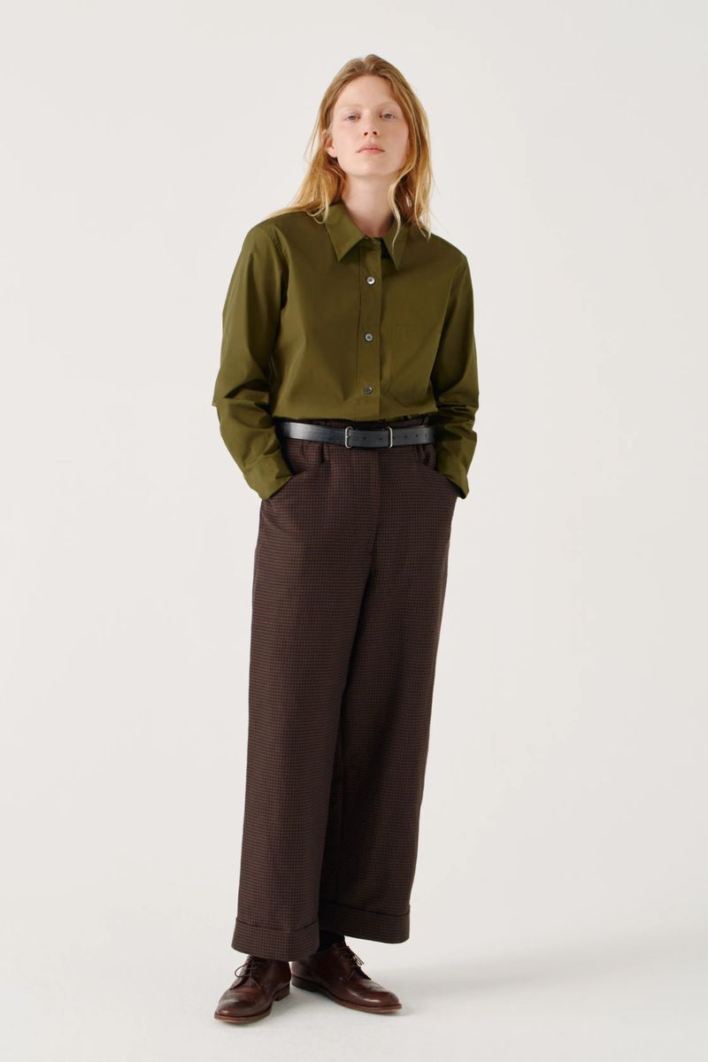 Full-length studio shot of a female model in an olive green shirt and brown patterned pants, standing against a white backdrop.