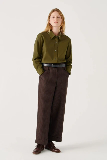 Full-length studio shot of a female model in an olive green shirt and brown patterned pants, standing against a white backdrop.