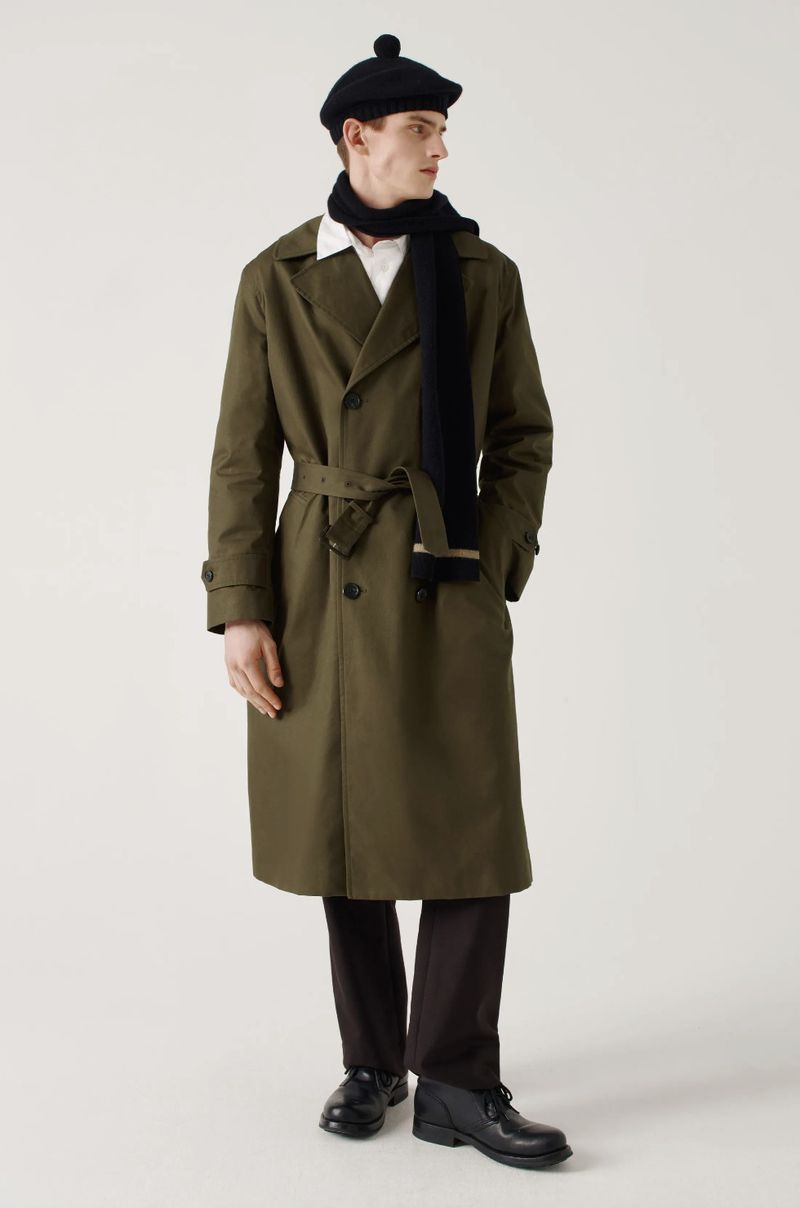 Man in an olive green trench coat, black scarf, and black beret standing in front of a plain off-white background in a studio.
