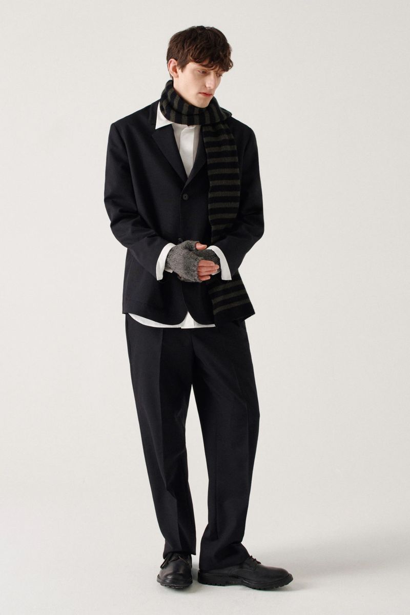 Man in a black suit, white shirt, and striped scarf stands in a brightly lit studio wearing fingerless gloves for a fashion forward pose.