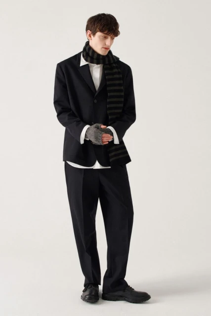 Man in a black suit, white shirt, and striped scarf stands in a brightly lit studio wearing fingerless gloves for a fashion forward pose.