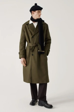 A man wearing an olive green trench coat, dark beret, and dark scarf against a plain white background for Margaret Howell F/W 26.