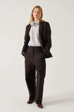 Model standing wearing dark brown blazer, white shirt with ruffled collar, and dark trousers against a white backdrop.