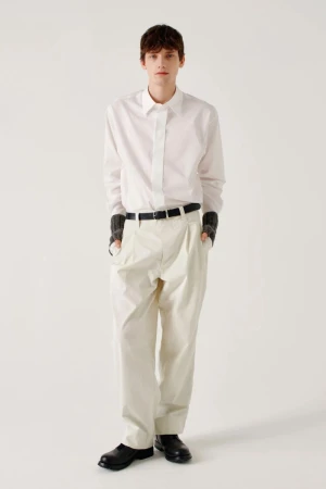 Model standing against a plain white background wearing a white long-sleeve button-up shirt, cream pleated trousers, and a black belt.