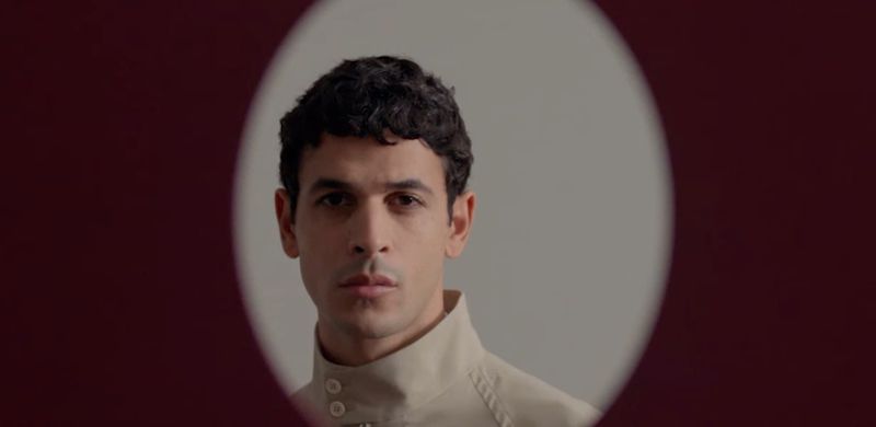 Portrait of a man wearing a high-collared cream shirt framed within an oval vignette against a deep burgundy backdrop
