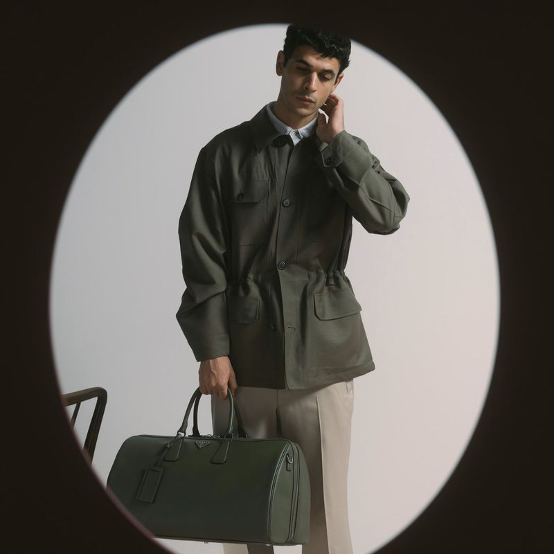 Adult male poses indoors, wearing olive green jacket and cream pants, holding a green leather duffel bag, shot through a circular frame.