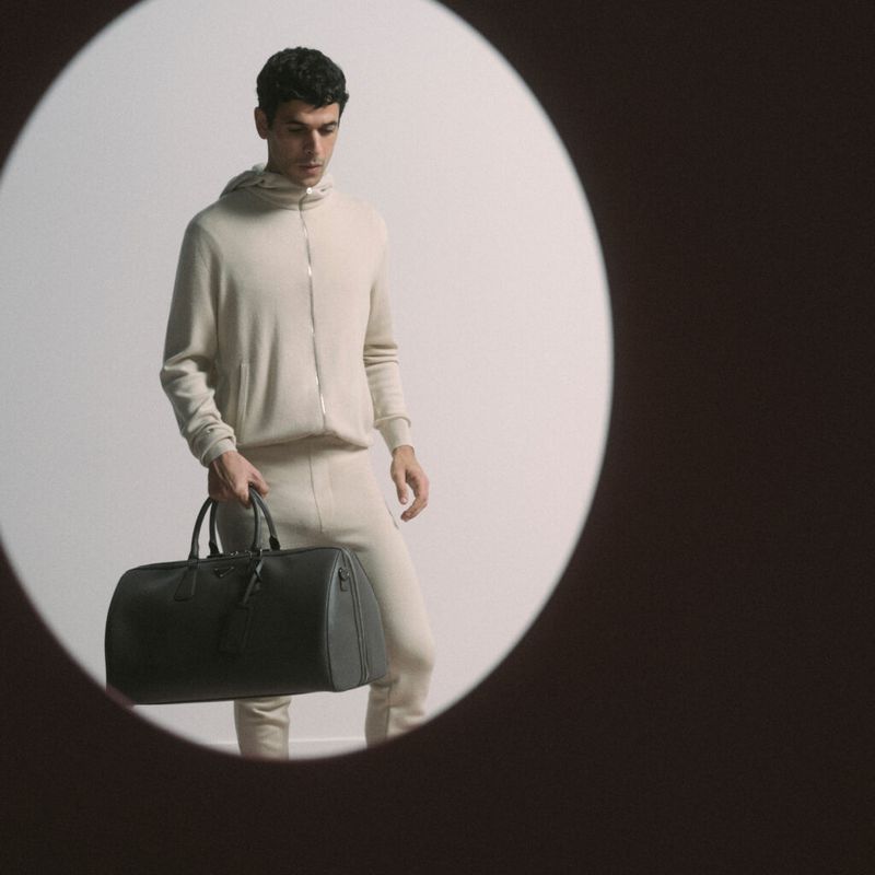 Man in a beige tracksuit holds a black duffel bag against a white backdrop illuminated by a spotlight, captured in a studio setting.