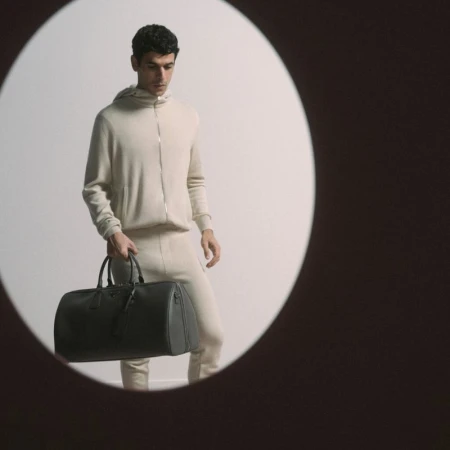 Man in a beige tracksuit holds a black duffel bag against a white backdrop illuminated by a spotlight, captured in a studio setting.