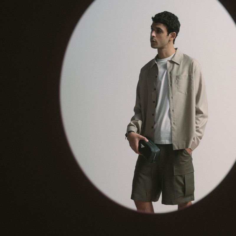 Studio shot of a young adult man wearing an open tan collared shirt and green shorts, holding a small dark green triangular case.