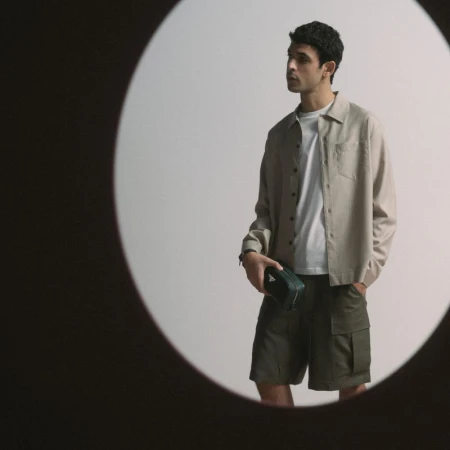 Studio shot of a young adult man wearing an open tan collared shirt and green shorts, holding a small dark green triangular case.