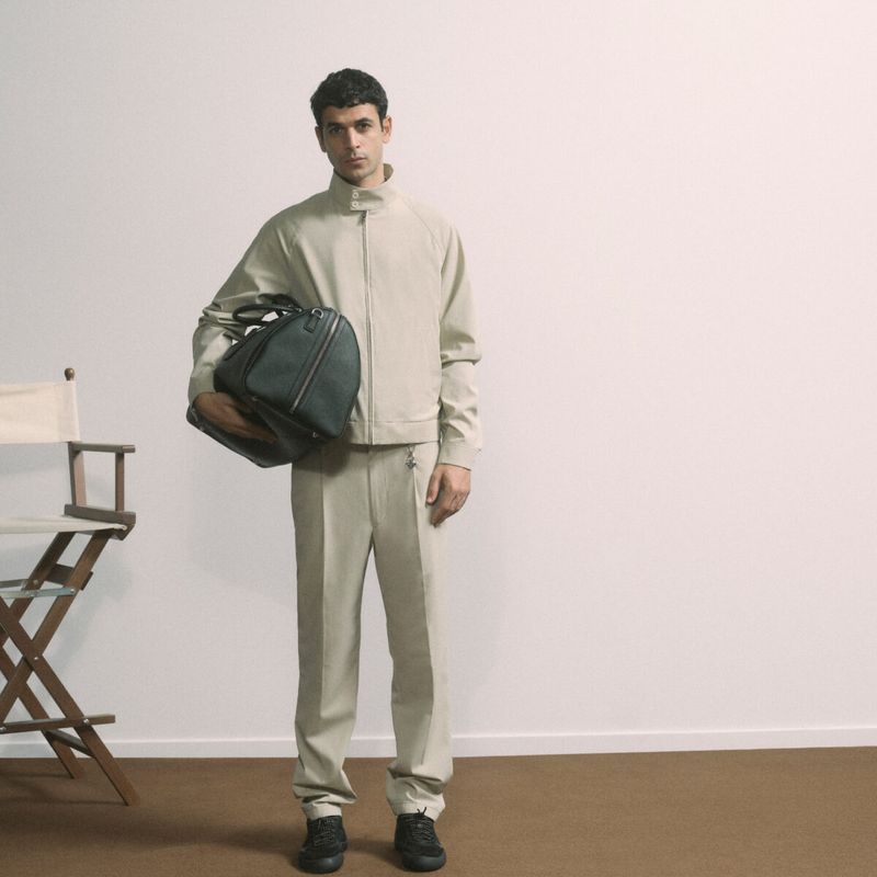 Man in a beige jacket and pants holding a dark green duffel bag, standing in front of a white wall next to a director's chair.