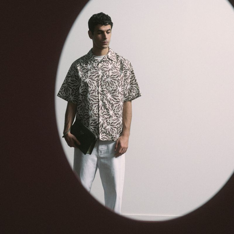 Man in patterned shirt and white pants holds a black briefcase, set against a light wall in a studio environment with creative framing.