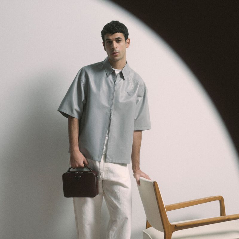 Model wearing a grey short-sleeve shirt and white trousers holding a brown leather handbag, standing beside a wooden chair.