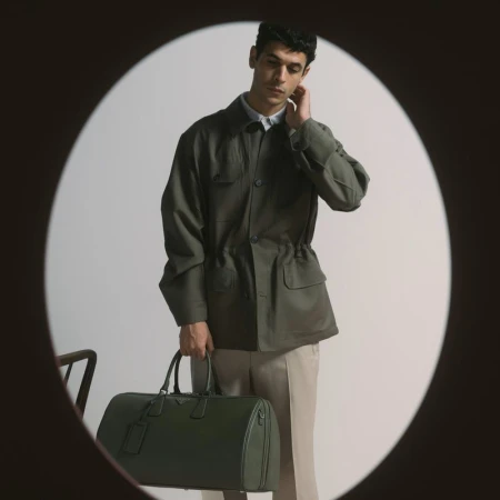 Man wearing a dark olive green jacket holding a dark green duffel bag viewed through a circular vignette frame.