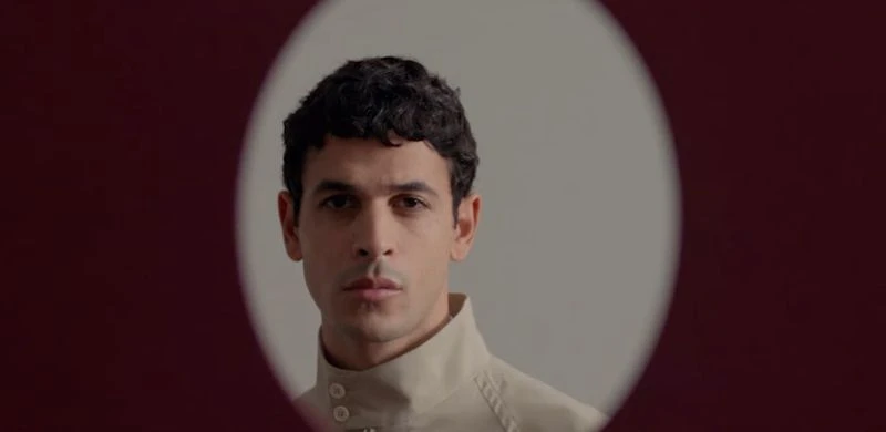 Portrait of a man wearing a high-collared cream shirt framed within an oval vignette against a deep burgundy backdrop