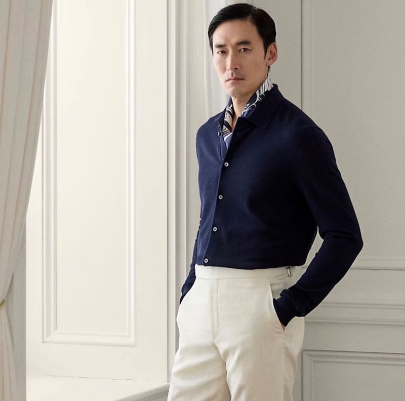 Portrait of an Asian man in a navy button-down shirt and white pants, standing near a bright window with white trim in a neutral setting.
