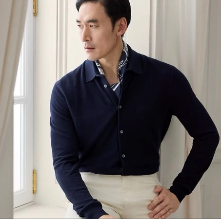 Asian man wearing a navy blue buttoned polo shirt and light trousers by a window with white curtains.