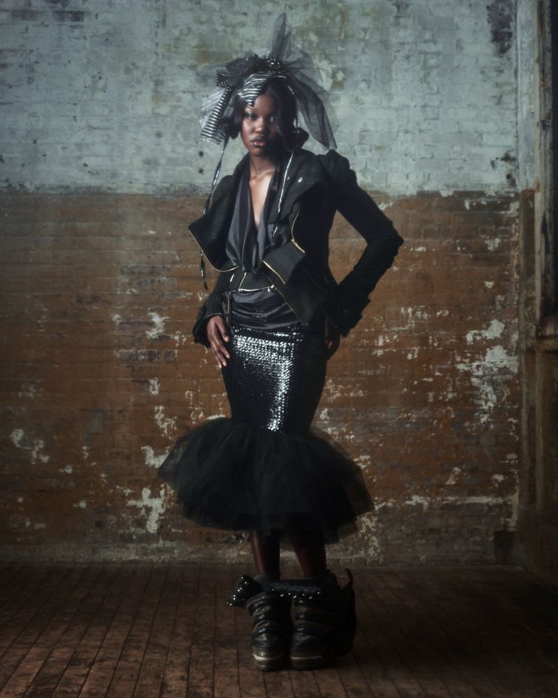 A woman with dark skin in a black jacket and black sequin skirt poses in a bare room with a brick wall and dark wood floor.