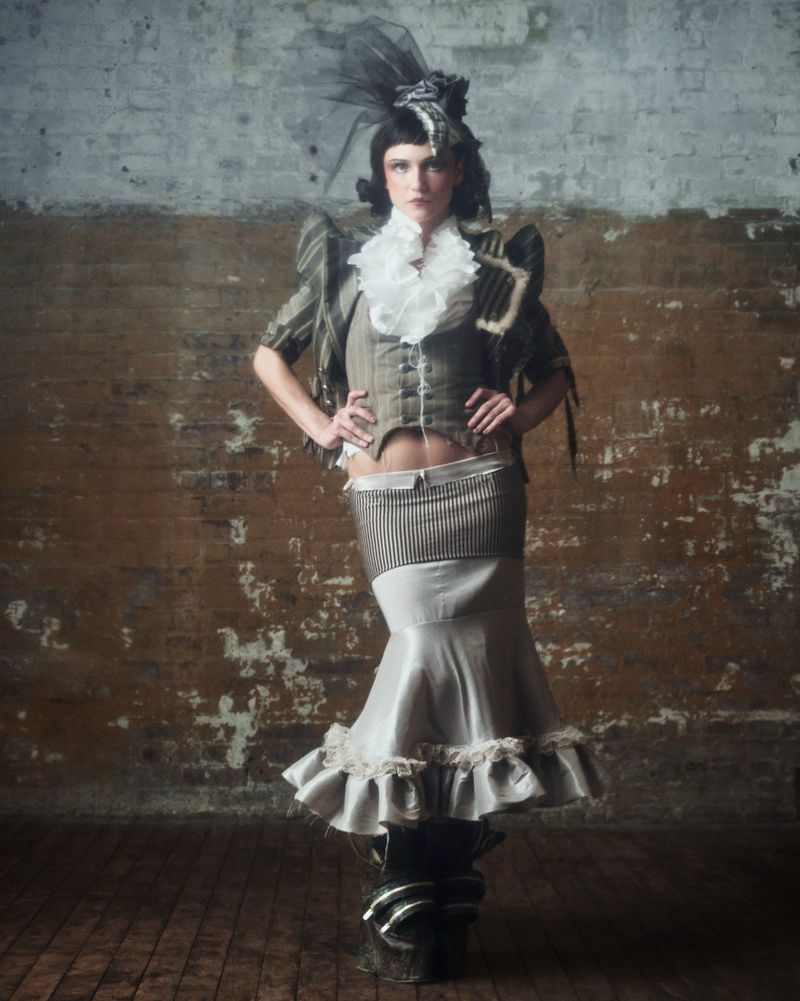 Elegant woman in an olive-green period costume posing against a distressed brick wall in a fashion photography style.