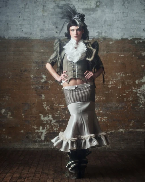 Elegant woman in an olive-green period costume posing against a distressed brick wall in a fashion photography style.